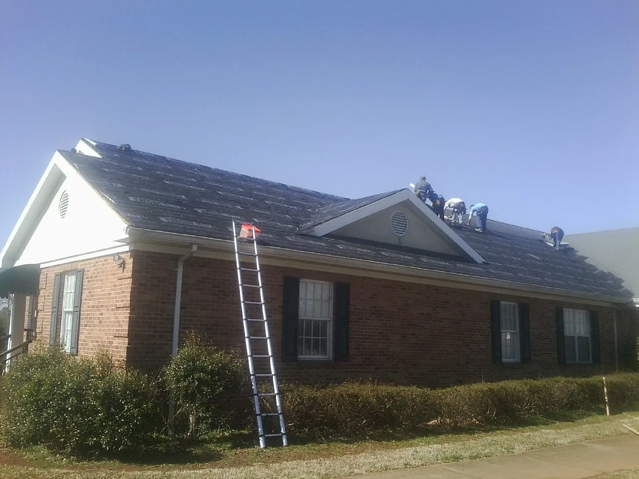 Crew tearing shingles off a brick ranch roof with a ladder set — Emergency Roof Repair work by our crew in Longwood
