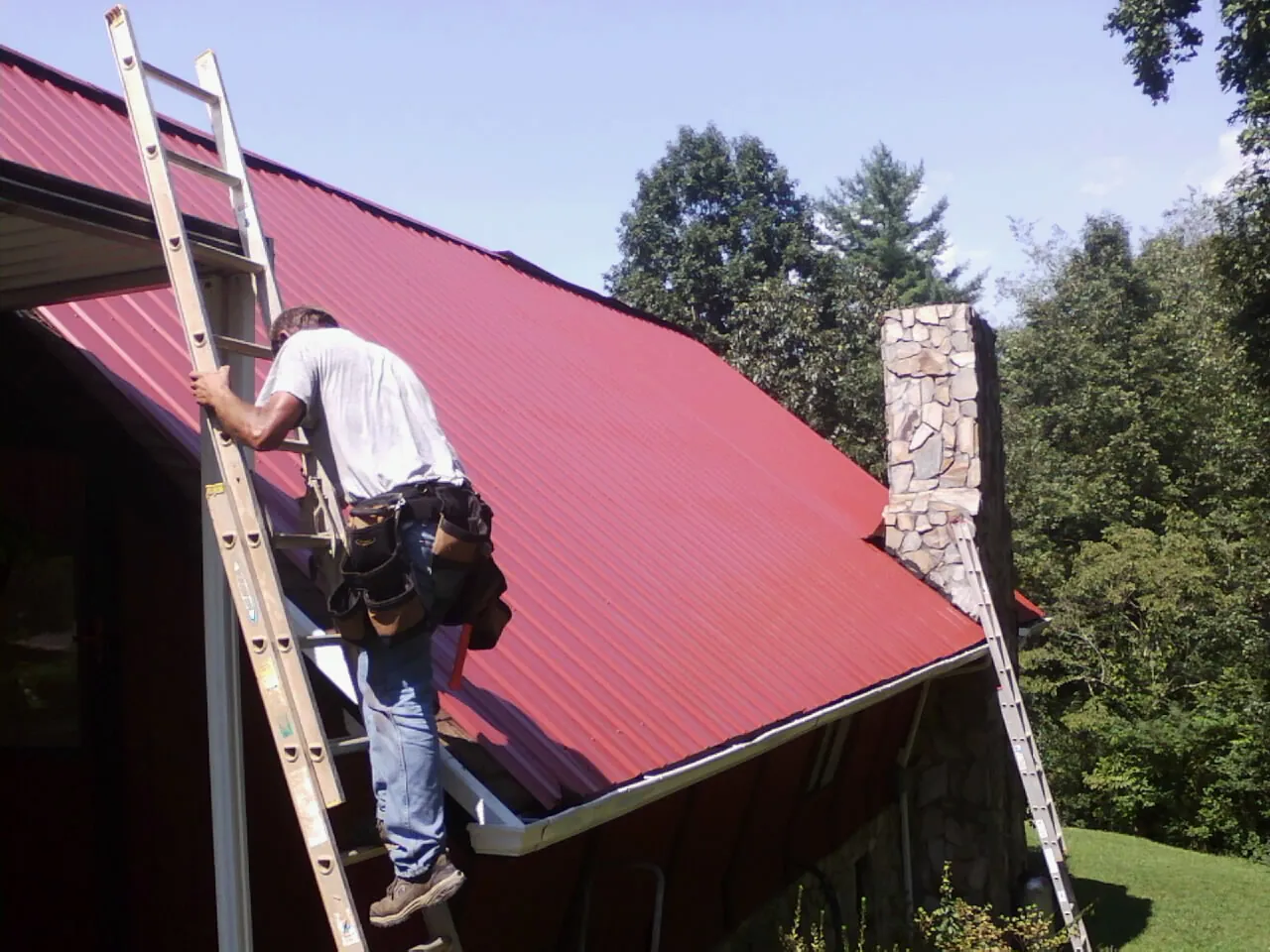 Roofer on a ladder beside a red metal roof and stone chimney — Soffit & Fascia Repair by Roofing Brothers in Longwood